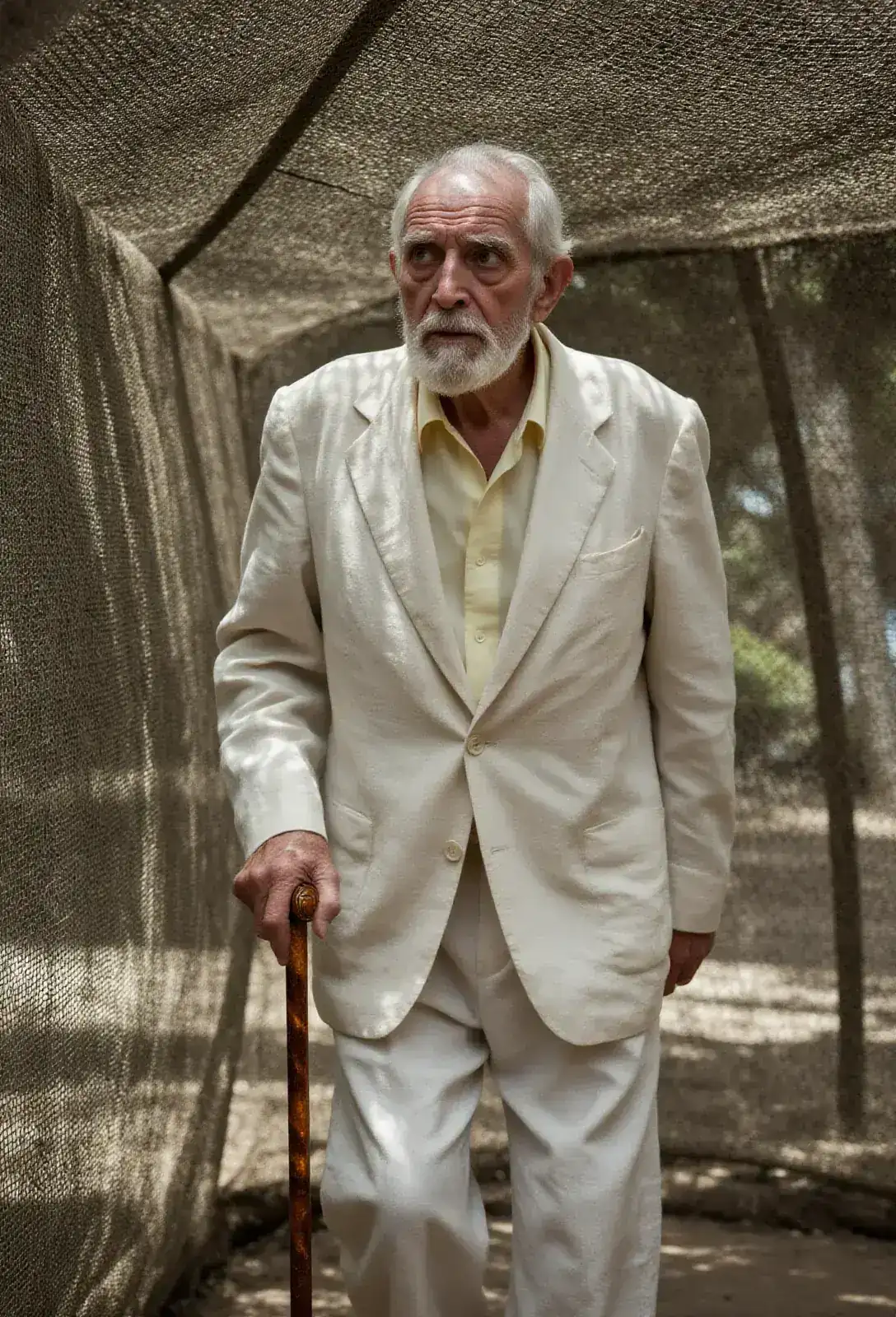 John Hammond walking under high netting, leaning on an amber-topped cane, white-suited and frail, with shafts of dappled sunlight through the netted dome.