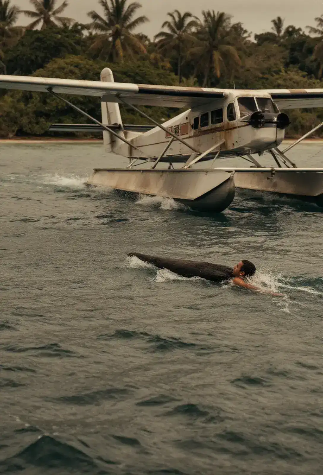 A pontoon plane banking back over flat tropical sea toward a man swimming alone, a long dark shape moving fast under the water below the plane.